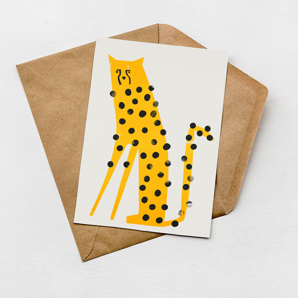 A greeting card with a yellow leopard design and black dots is placed on a brown envelope against a white background.