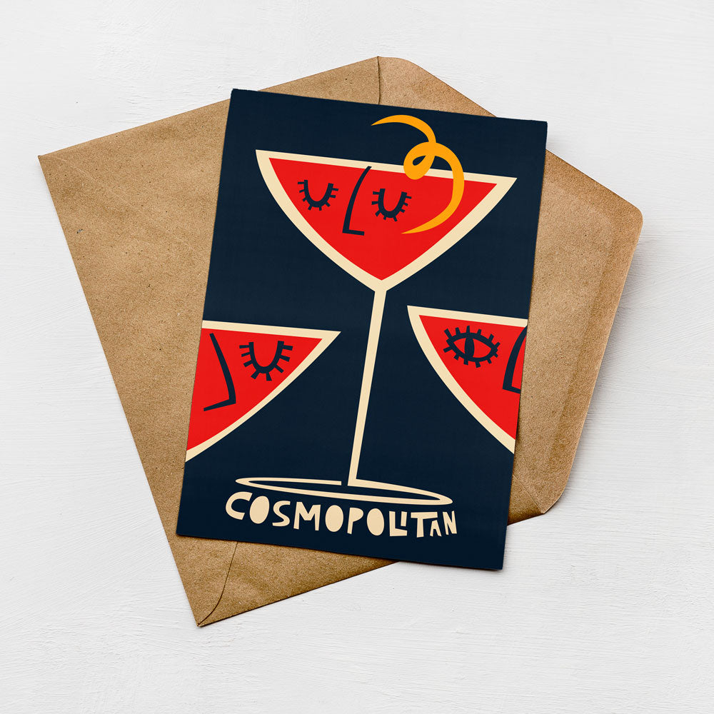 A greeting card with a stylized illustration of a martini glass and the word "cosmopolitan" printed on it, placed on a brown envelope against a white background.