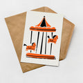 A card with an illustration of a carousel featuring horses in orange and black colors, placed on top of a brown envelope.