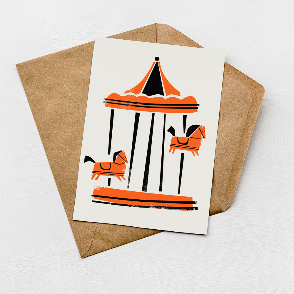 A card with an illustration of a carousel featuring horses in orange and black colors, placed on top of a brown envelope.