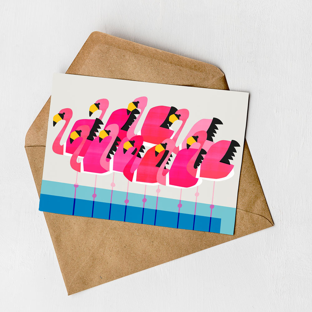 A greeting card featuring a vibrant illustration of pink flamingos with yellow beaks and feet, set against a blue background with a white border.