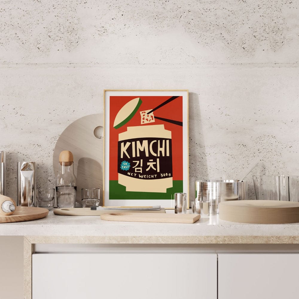 Kimchi print framed on a kitchen shelf with stone backsplash and assorted glassware.