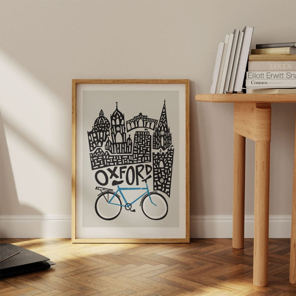 The Oxford print leaning casually against a wall beside a desk, its architectural skyline rising above a vintage-style blue bicycle in crisp, graphic lines.