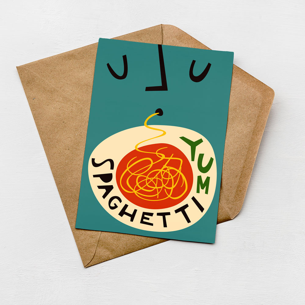 A greeting card with a colorful illustration of spaghetti and the words "YUM SPAGHETTI" printed on it, placed on a brown envelope against a white background.