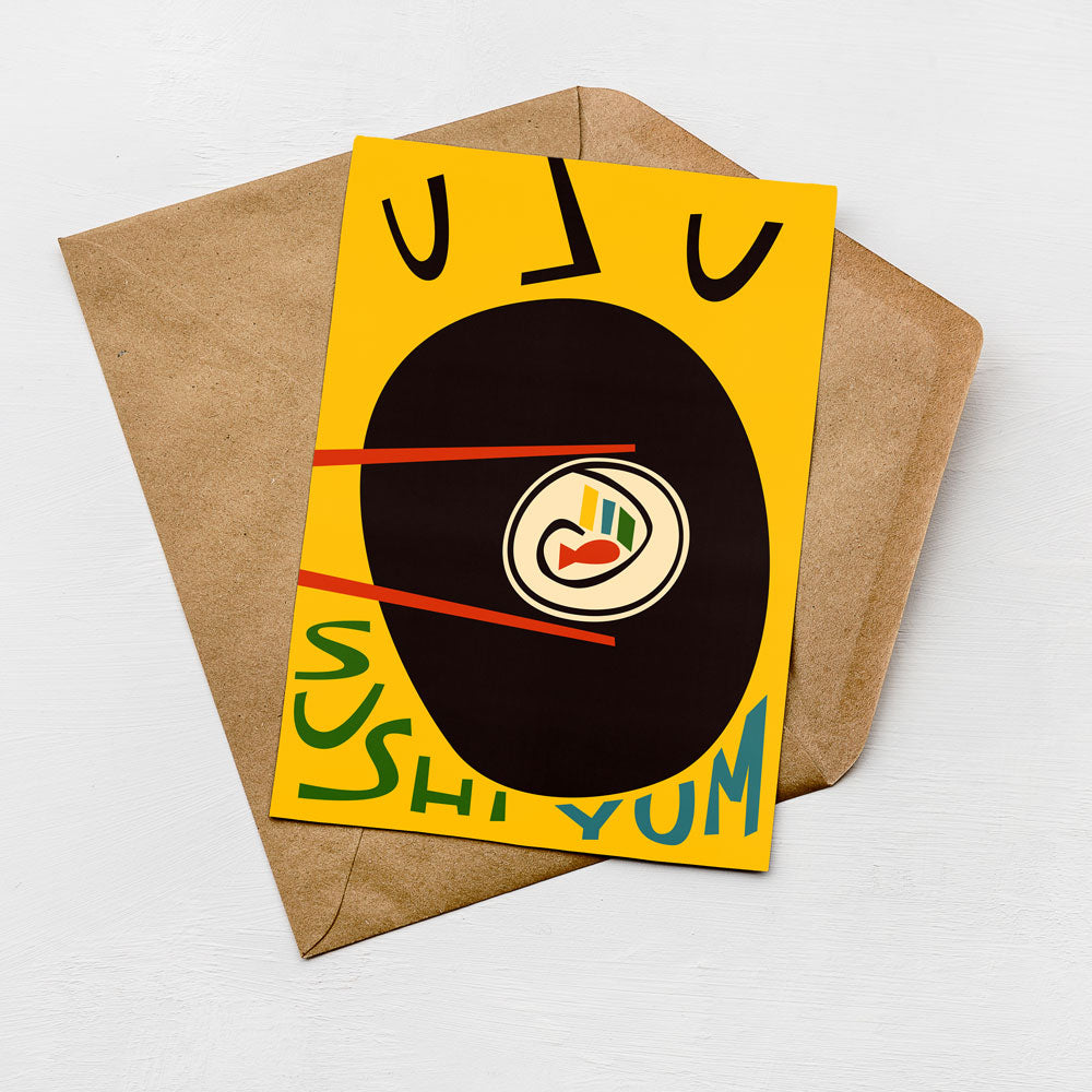 A yellow card with a black and white illustration of a sushi dish, along with the text "SUSHI YUM" written on it, placed on a brown envelope.
