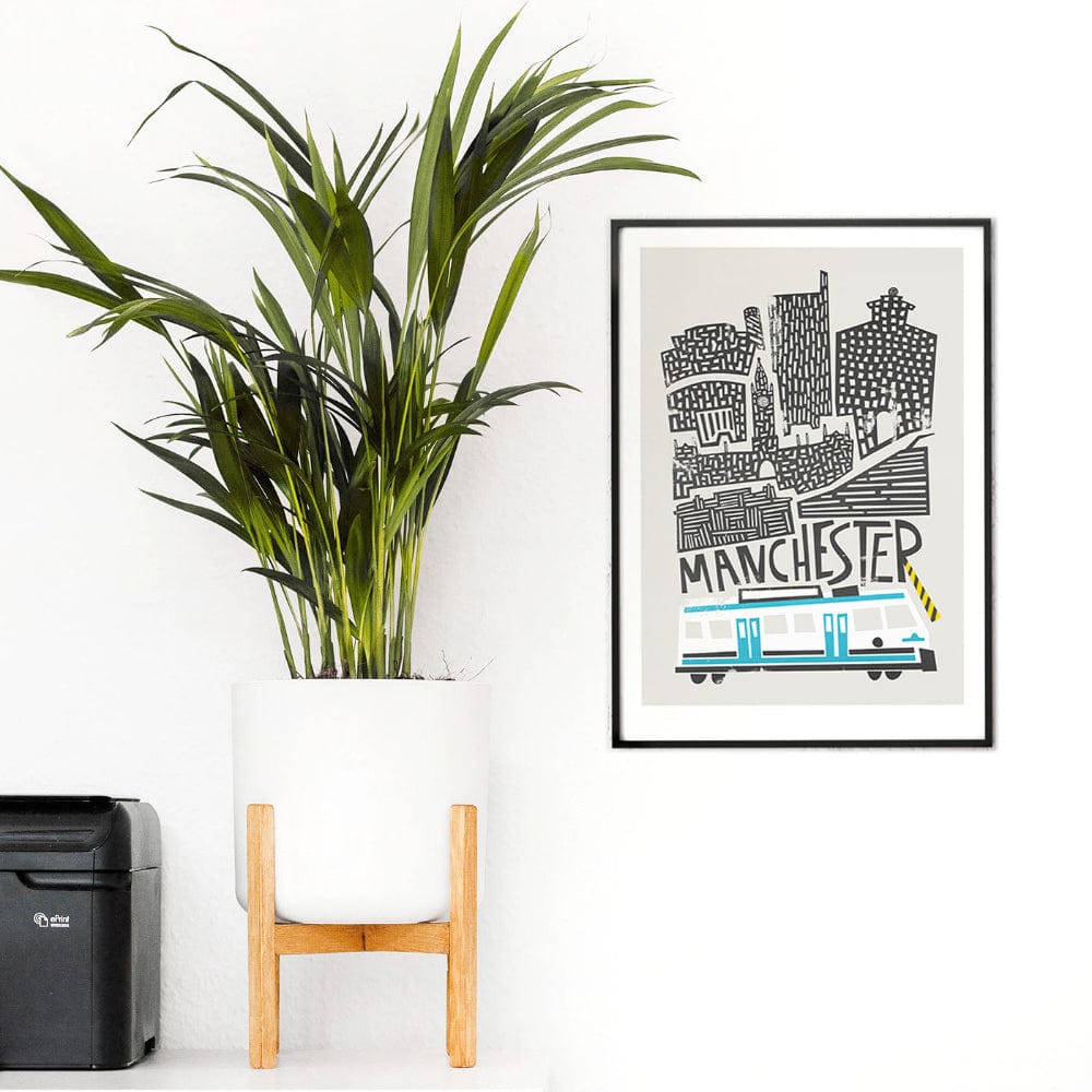 Framed Manchester city print hung on a minimalist white wall beside a plant, capturing the sleek contrast between graphic black lines and soft blue tram details.