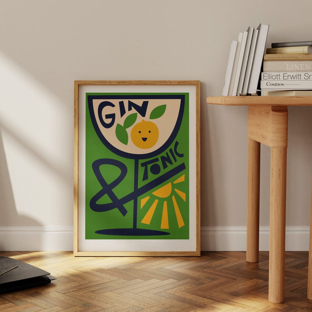 Gin & Tonic print propped on the floor beside a wooden desk and books, adding personality to a calm, casual interior.