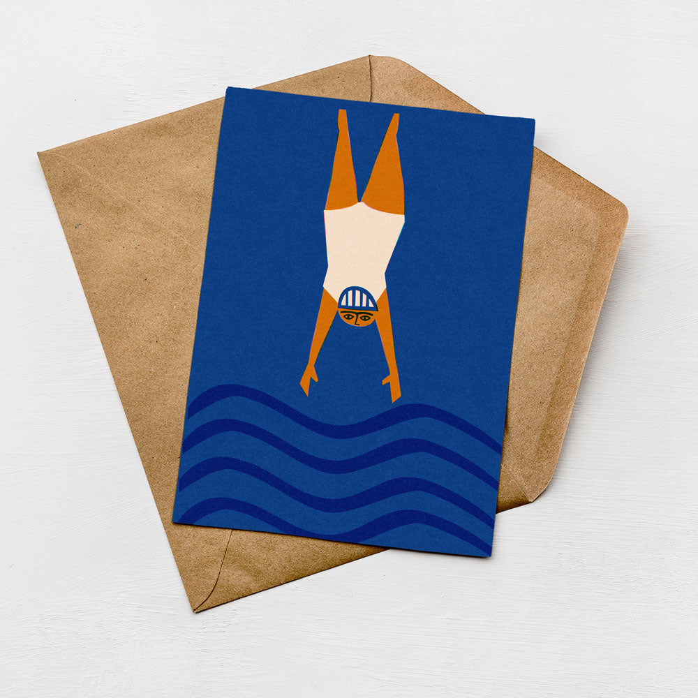 A greeting card with a blue background and a swimming illustration is placed on top of a brown envelope.