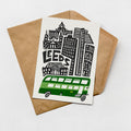 A greeting card featuring an illustration of a city skyline with a green double-decker bus in the foreground.
