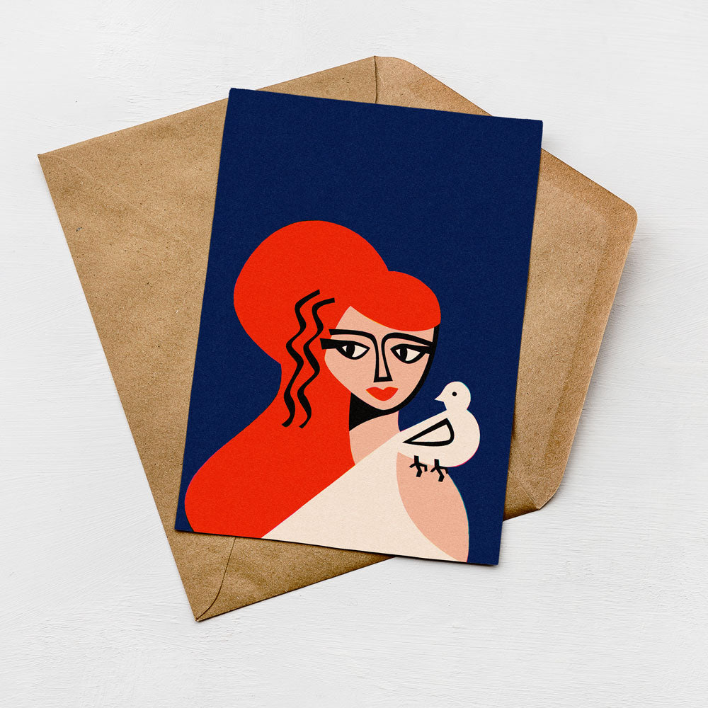 a card with a stylized illustration of a woman with red hair and a bird. The card is placed on top of a brown envelope, which is also on a white surface.