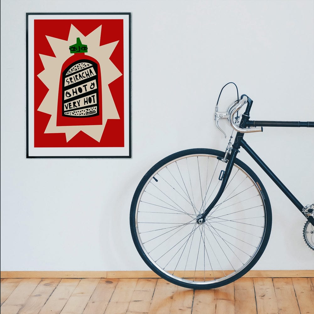 Sriracha hot sauce artwork framed on a clean white wall beside a bike and wooden floor.