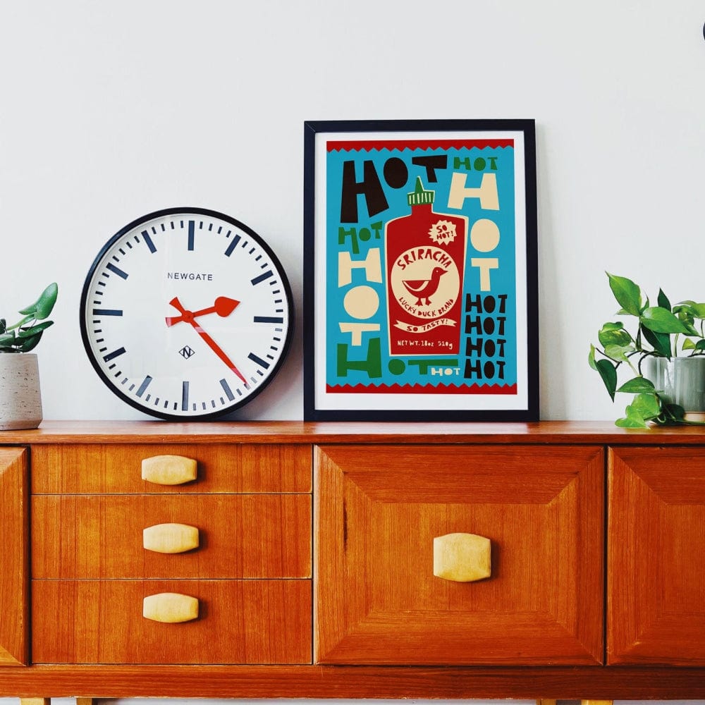 Sriracha duck print framed above a mid-century sideboard with a large clock and small plant.