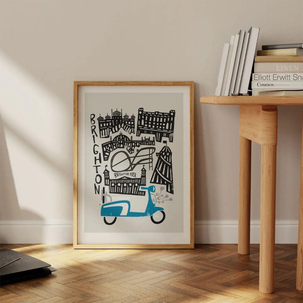 Framed Brighton city art print resting on a wooden floor beneath a desk, bringing a playful yet stylish coastal charm to the space.