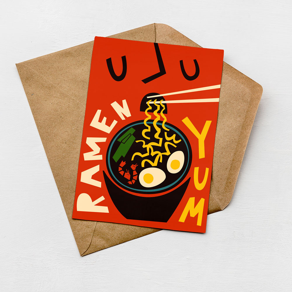 A vibrant red greeting card with a playful illustration of a bowl of ramen noodles and the text "RAMEN YUM" on it, placed on a brown envelope against a white background.