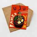 A vibrant red greeting card with a playful illustration of a bowl of ramen noodles and the text "RAMEN YUM" on it, placed on a brown envelope against a white background.