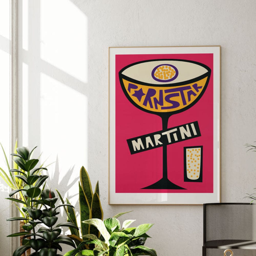 The vibrant Pornstar Martini print pops against a white wall, framed in black and surrounded by lush green plants in a light-filled modern room.