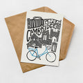 A greeting card featuring an illustration of a cityscape with a bicycle, placed on a brown envelope against a white background.